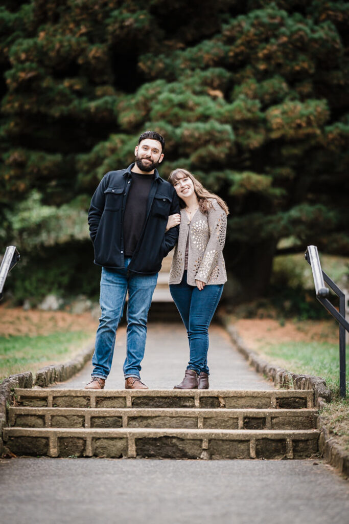 Engaged couple on stone steps with evergreen trees at Deep Cut Gardens - Middletown NJ engagement photographer Alex Kaplan