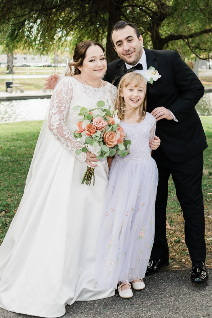 Happy family portrait with bride, groom, and flower girl under willow tree.