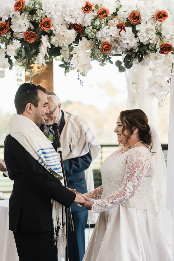Bride and groom holding hands under chuppah during Jewish ceremony.