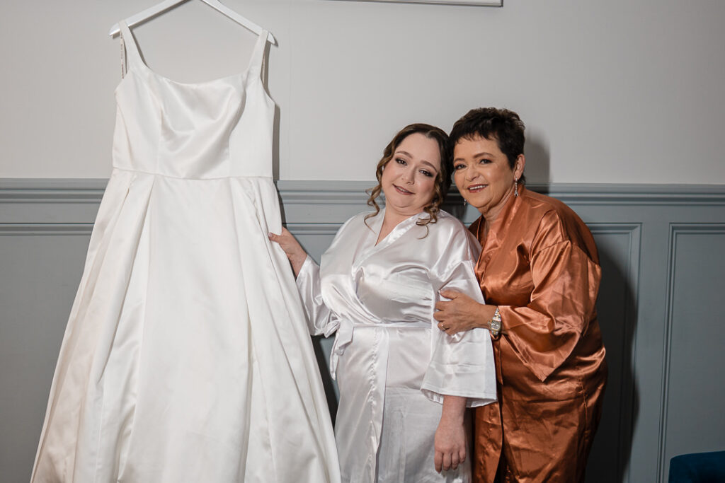Bride and mother in robes admiring white satin wedding dress before ceremony.
