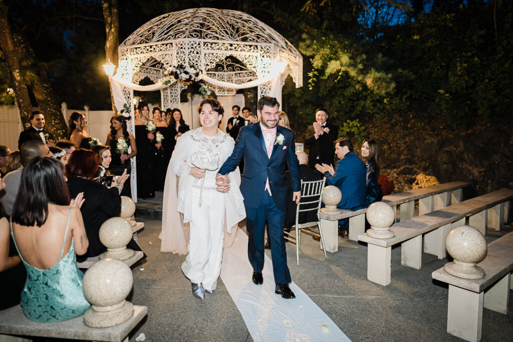 Newlyweds walking down aisle as guests applaud at illuminated gazebo Cortlandt Manor NY