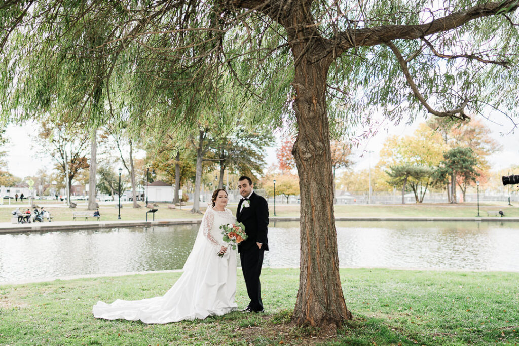 Bride and groom standing under large willow tree by pond in Lincoln Park.