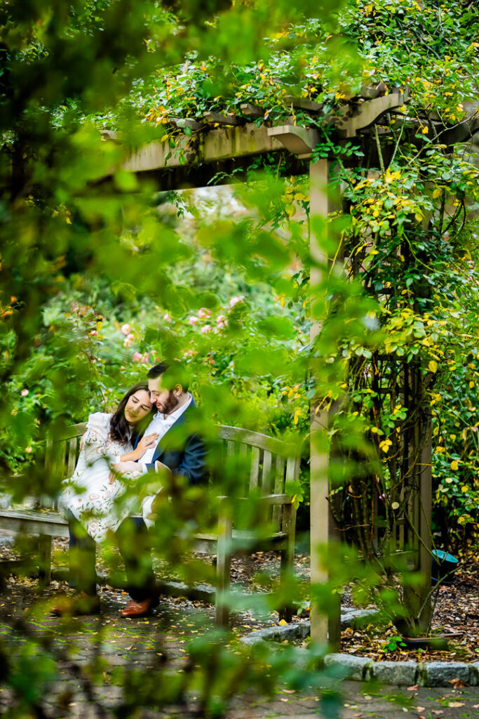 Engaged couple sitting close on a garden bench framed through green foliage