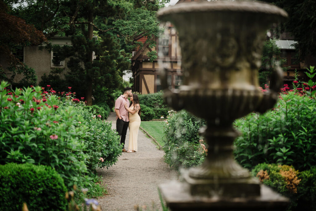 Garden engagement session with couple walking among roses photographed through ornate fountain for creative composition