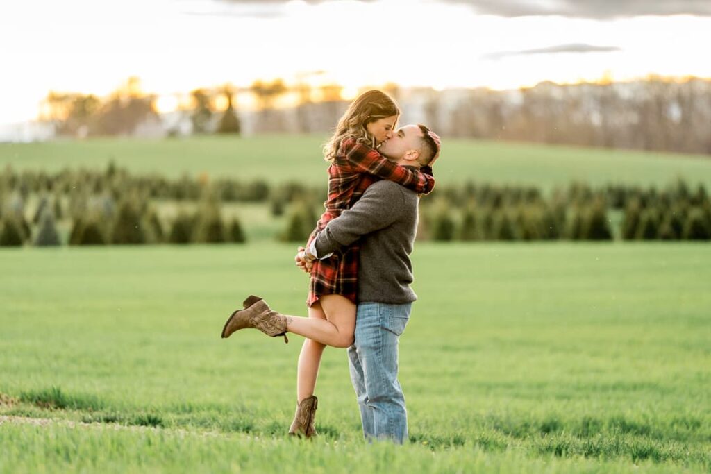 Playful golden hour engagement photo at tree farm with couple kissing during sunset with warm backlighting