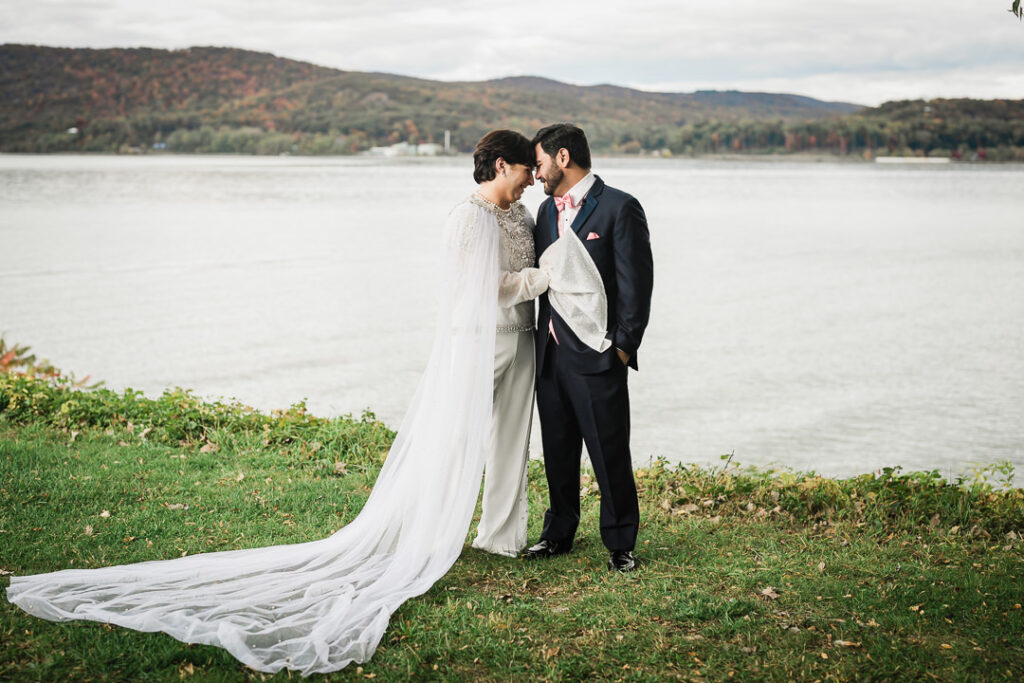 Couple standing by Hudson River with cathedral veil and autumn mountains, romantic Hudson Valley wedding portrait