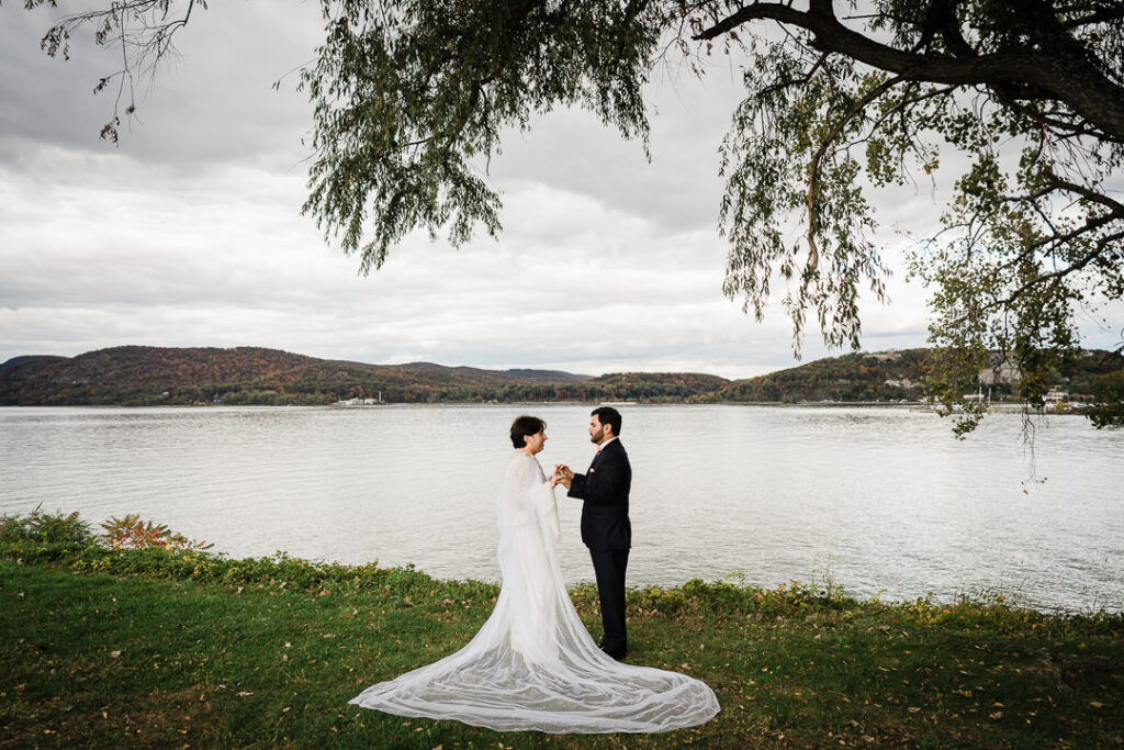 Wide landscape of couple holding hands beneath willow by Hudson River with autumn foliage