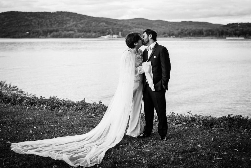 Black-and-white portrait of couple kissing by Hudson River with veil and mountain landscape, artistic Peekskill wedding photo