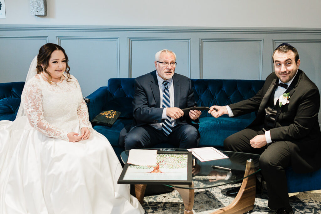 Bride and groom with rabbi during ketubah signing ceremony on blue velvet couch.