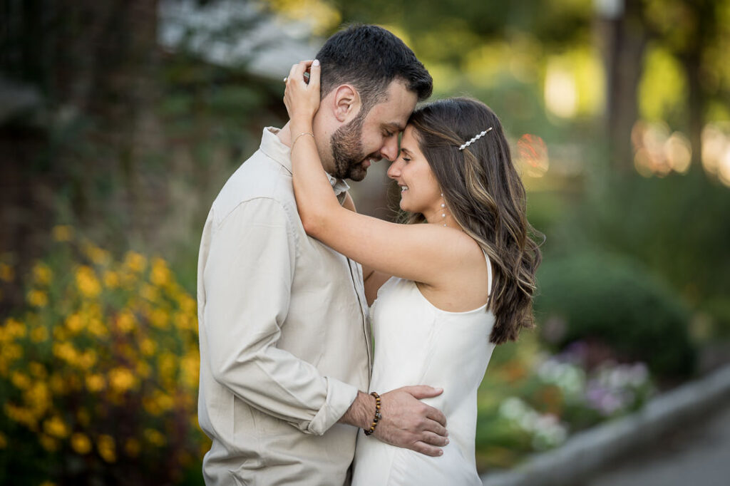 Engaged couple standing forehead to forehead beside a flower garden at golden hour