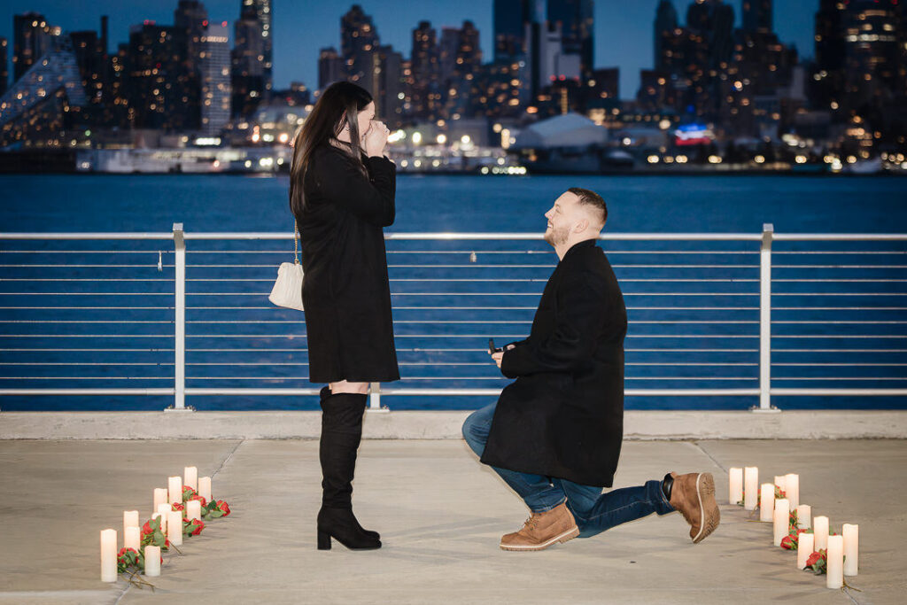 Emotional surprise proposal moment man on one knee with candles and roses at Hoboken Pier A waterfront with NYC skyline at twilight