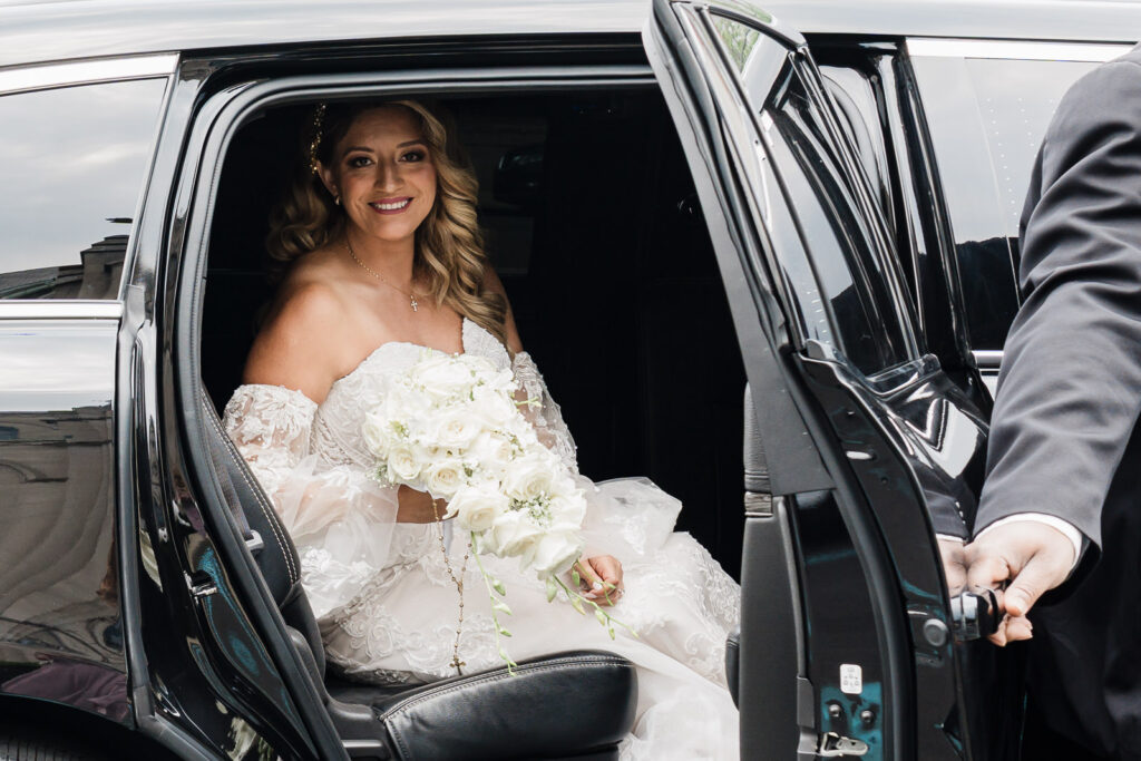 Bride in off-shoulder lace wedding dress with white rose bouquet arriving at church from limousine photographed by Alex Kaplan Photography