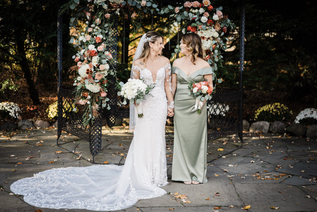 Bride and maid of honor in sage dress holding hands at ceremony gazebo with fall floral arch