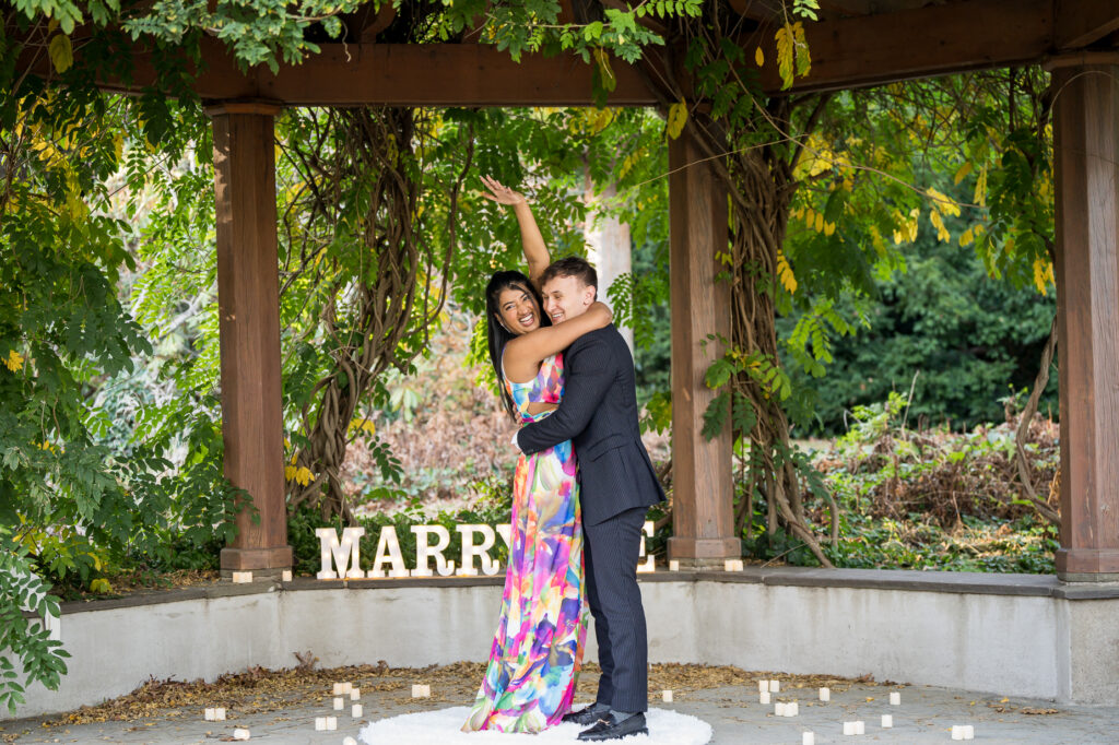 Engaged couple sharing kiss under vine-covered pergola surrounded by autumn garden colors