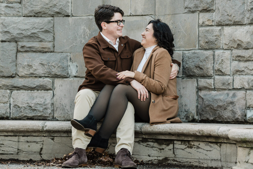 Couple sitting on stone wall laughing together during Verona Park engagement shoot with Alex Kaplan
