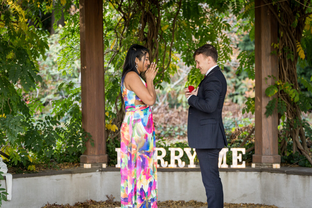 Man presenting ring box to emotional girlfriend at fall garden pergola with Marry Me sign and lush greenery Northern New Jersey