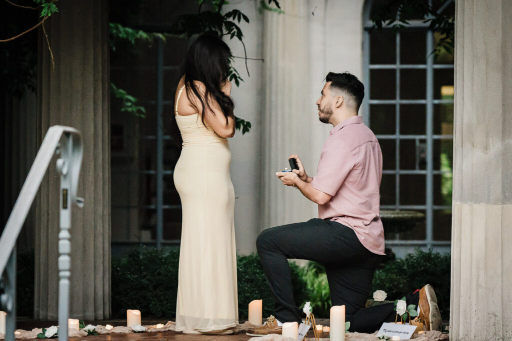 Surprise proposal moment man on one knee with ring at elegant garden setting with candles and white roses NJ