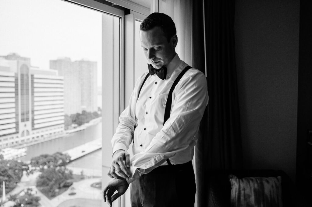 Black and white photo of groom adjusting cufflinks by window in tuxedo shirt and suspenders at Westin Newport Jersey City by Alex Kaplan Photography