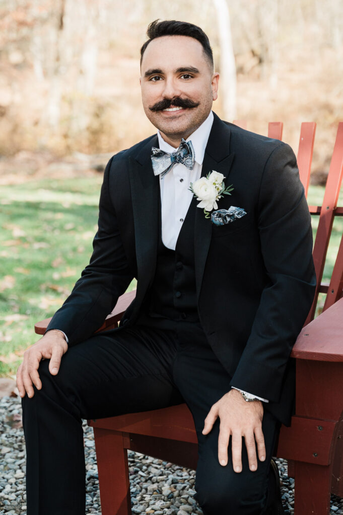 Groom in black tuxedo and patterned bow tie sitting outdoors for fall wedding portrait