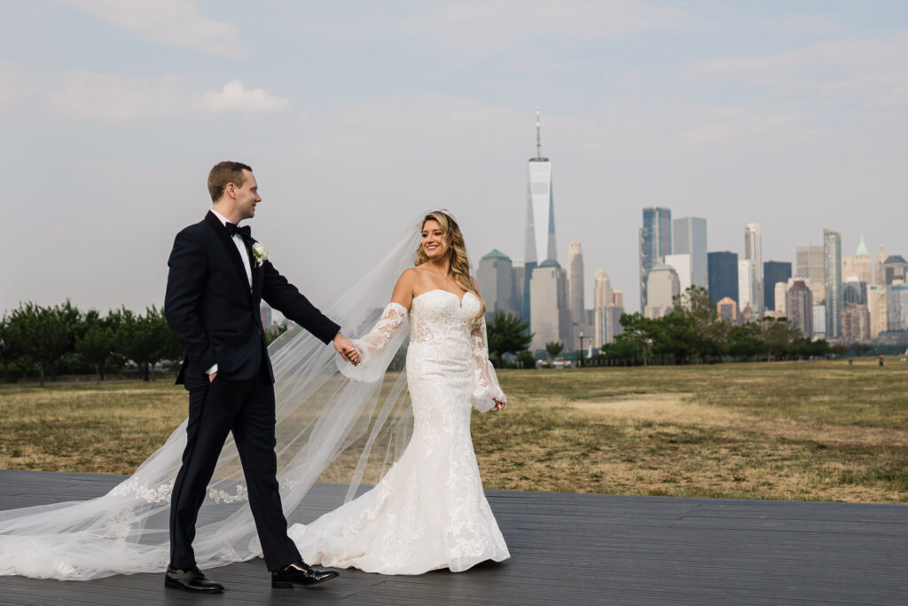 Bride holding veil with groom and NYC Manhattan skyline at Liberty House Liberty State Park wedding by Alex Kaplan Photography