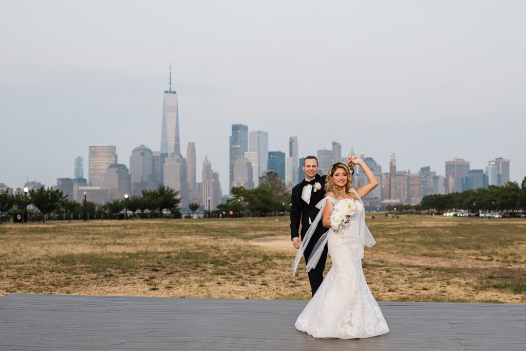 Bride posing with flowing veil and white rose bouquet on boardwalk with NYC Manhattan skyline at Liberty State Park Jersey City wedding by Alex Kaplan Photography