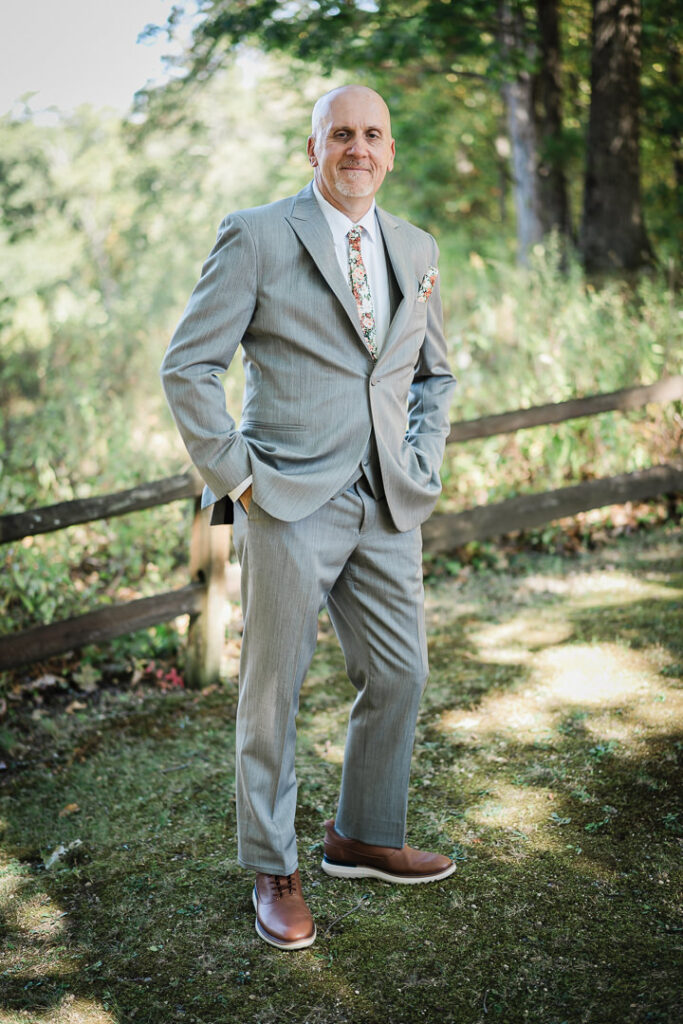 Groom in gray suit with floral tie poses outdoors at Mohawk House wedding venue in Sparta NJ photographed by Alex Kaplan