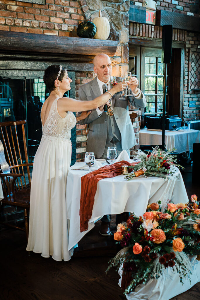 Couple toasts at sweetheart table with fireplace backdrop at Mohawk House Sparta NJ by Alex Kaplan Photography