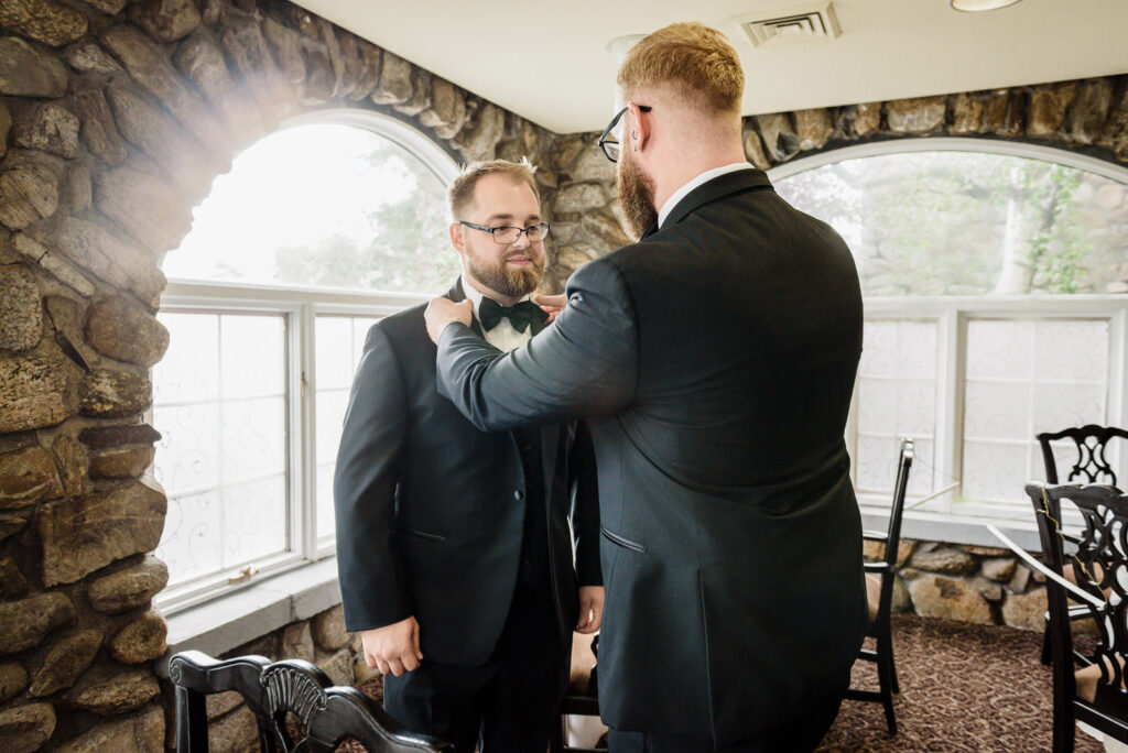 Groomsmen helping groom with bow tie in stone-walled prep room at Ramsey Country Club by Alex Kaplan Photography