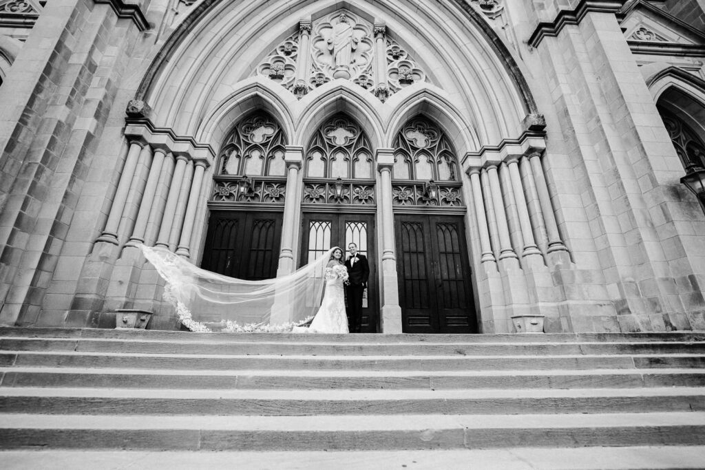 Bride and groom on steps of St. Henry's Church with cathedral veil and Gothic architecture exterior in Bayonne NJ by Alex Kaplan Photography