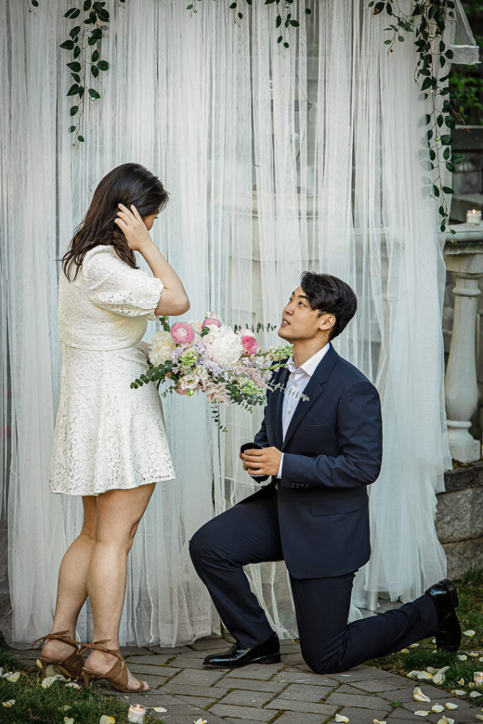 Man proposing on one knee to emotional bride-to-be at elegant outdoor setup with white drapes and flowers