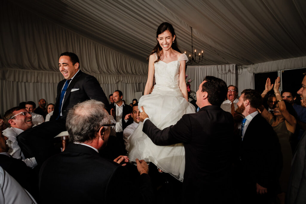 Radiant bride laughing joyfully while lifted high on chair during energetic Hora dance celebration