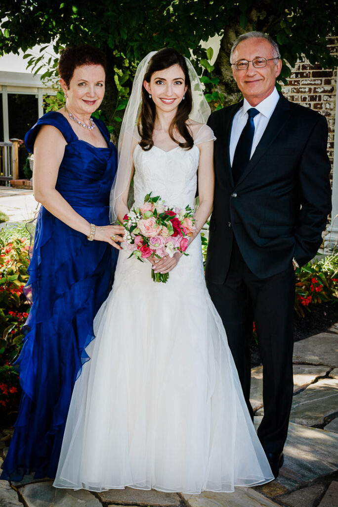 Bride with parents in formal attire on stone pathway with lush gardens at Hamilton Farm