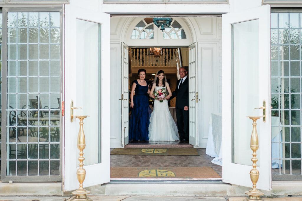 Bride standing with parents in elegant doorway framed by white columns and brass candlesticks at Hamilton Farm