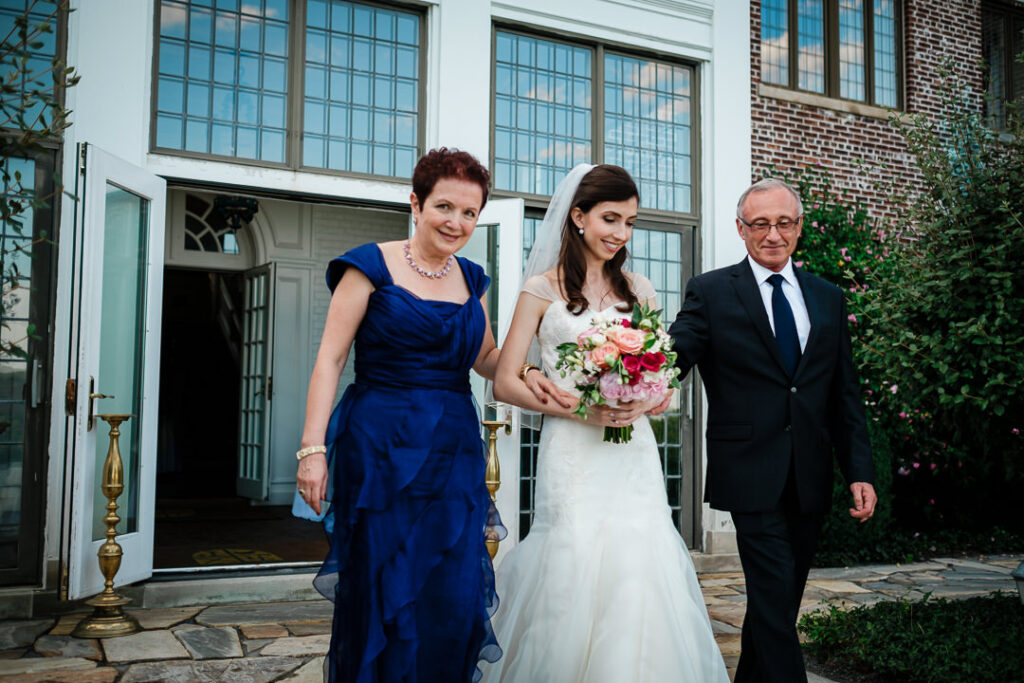 Bride walking down aisle with mother in royal blue dress and father toward outdoor Jewish ceremony