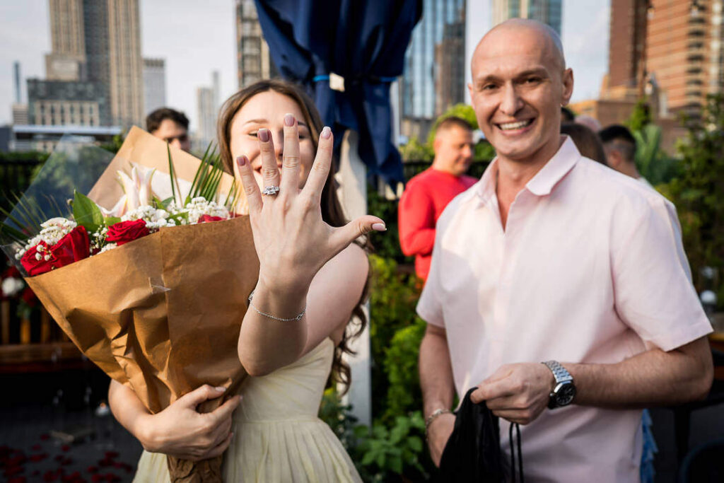 Excited bride shows off engagement ring to camera holding flower bouquet with groom and guests celebrating, Alex Kaplan Photography Northern New Jersey