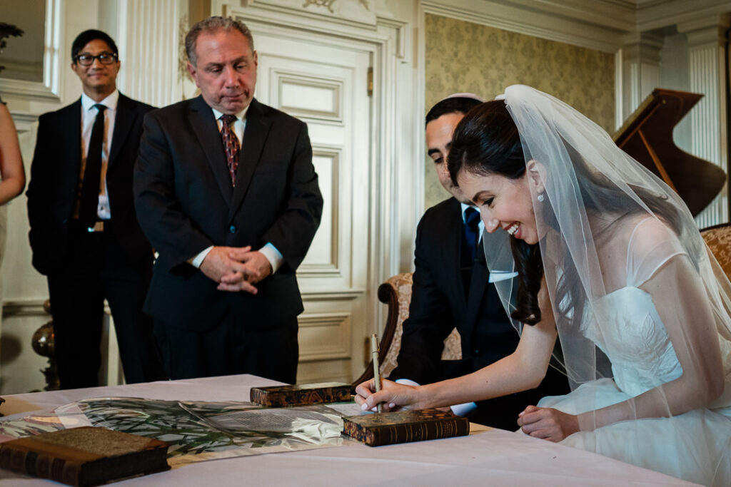 Bride signing ornate Ketubah marriage contract with rabbi and groom watching in elegant interior room