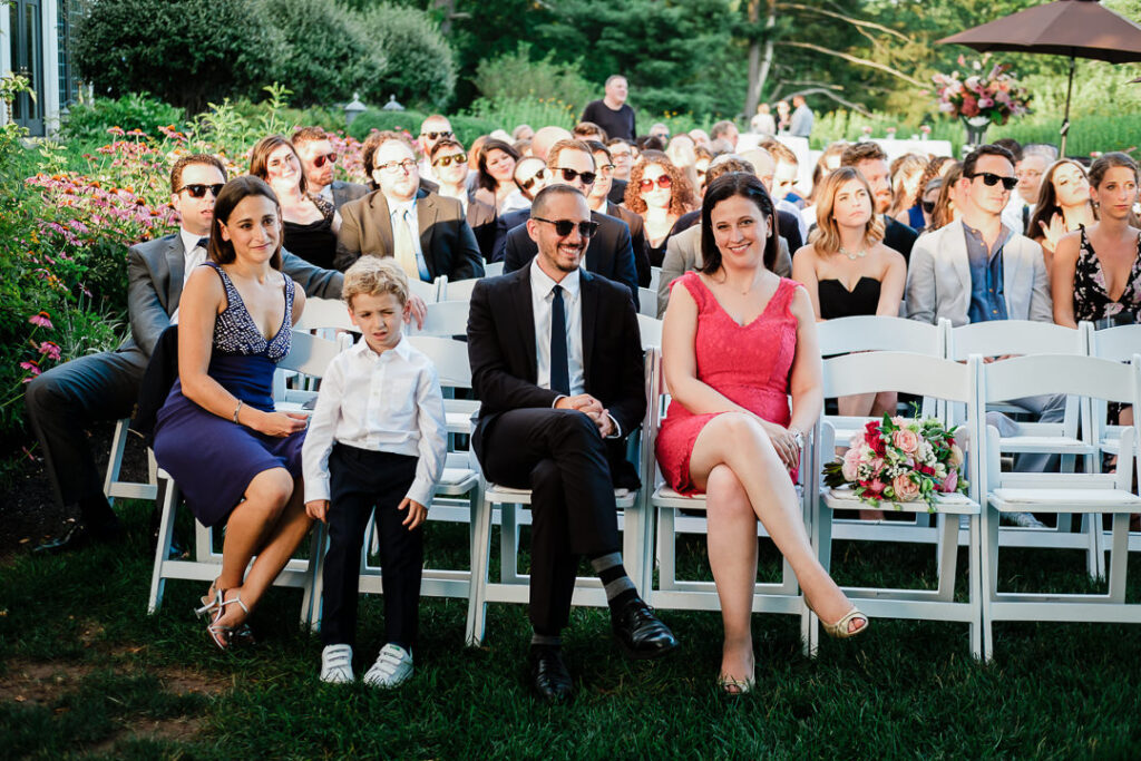 Wedding guests seated in white chairs watching Jewish ceremony with young ring bearer and flower arrangements