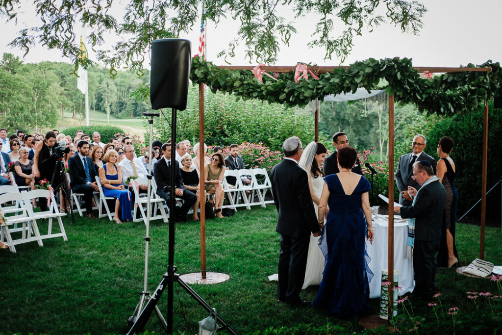 Bride and groom walking toward wooden chuppah adorned with pink florals as guests watch Jewish wedding ceremony