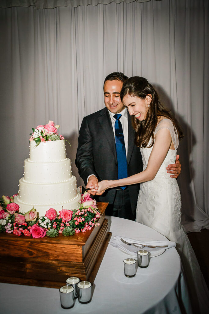 Happy couple cutting four-tier wedding cake together surrounded by pink roses and pearl details