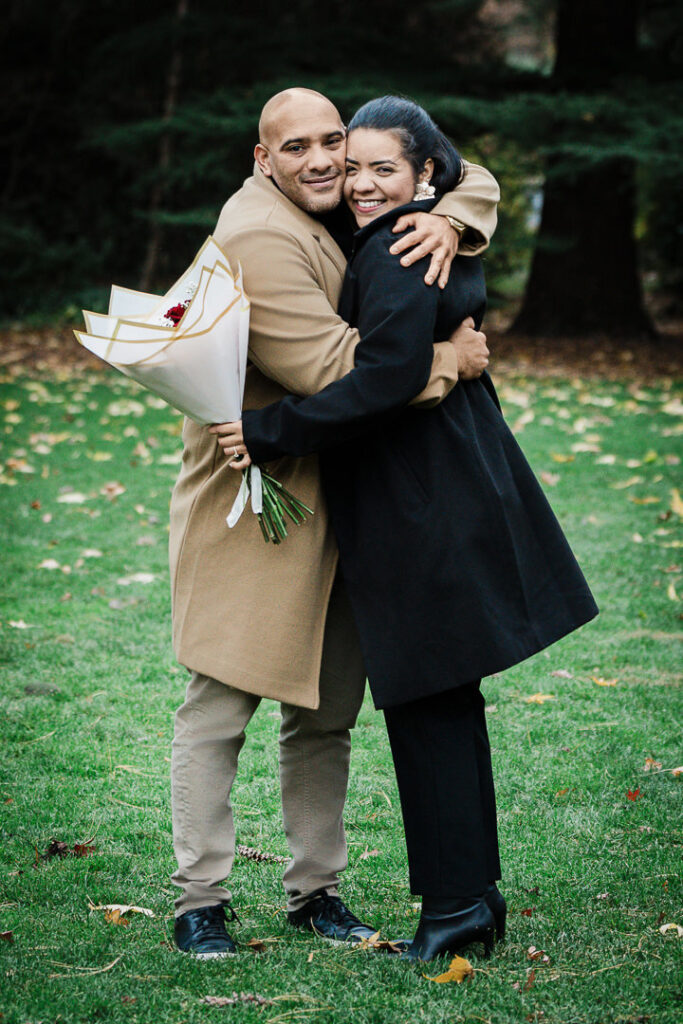 Engaged couple embraces on Van Vleck garden lawn with red roses bouquet, fall engagement session Northern New Jersey photographer