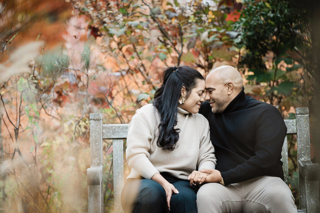 Newly engaged couple in intimate embrace on Van Vleck garden bench with autumn colors backdrop, professional engagement photography Montclair NJ