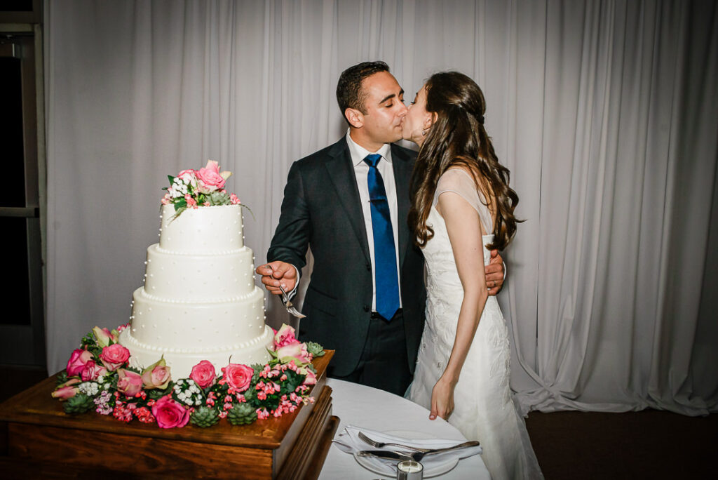 Newlyweds sharing sweet kiss after cutting elegant four-tier wedding cake at Hamilton Farm reception