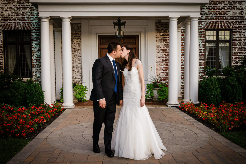 Romantic kiss between couple framed by white columns and wooden doors at Hamilton Farm grand entrance