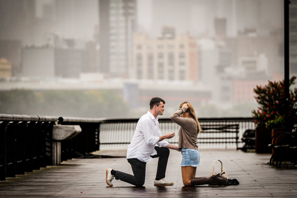 Happy engaged couple embracing on Pier 13 boardwalk in Hoboken NJ with misty Manhattan skyline across Hudson River