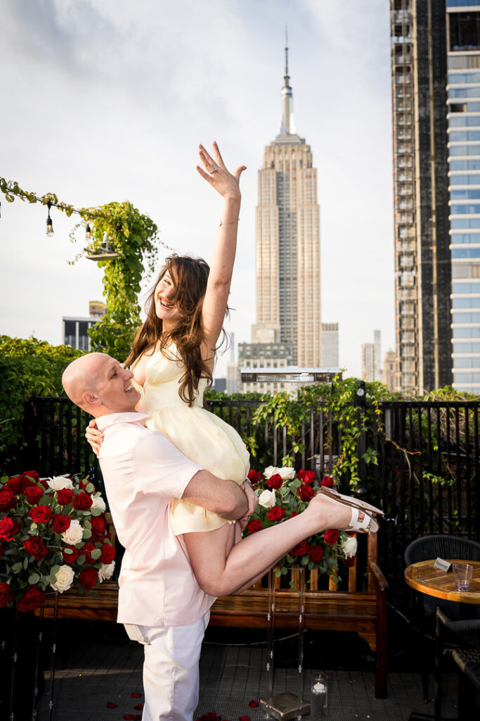 Engaged couple celebrates with joyful pose on rooftop with Empire State Building prominently featured, professional proposal photographer NYC area