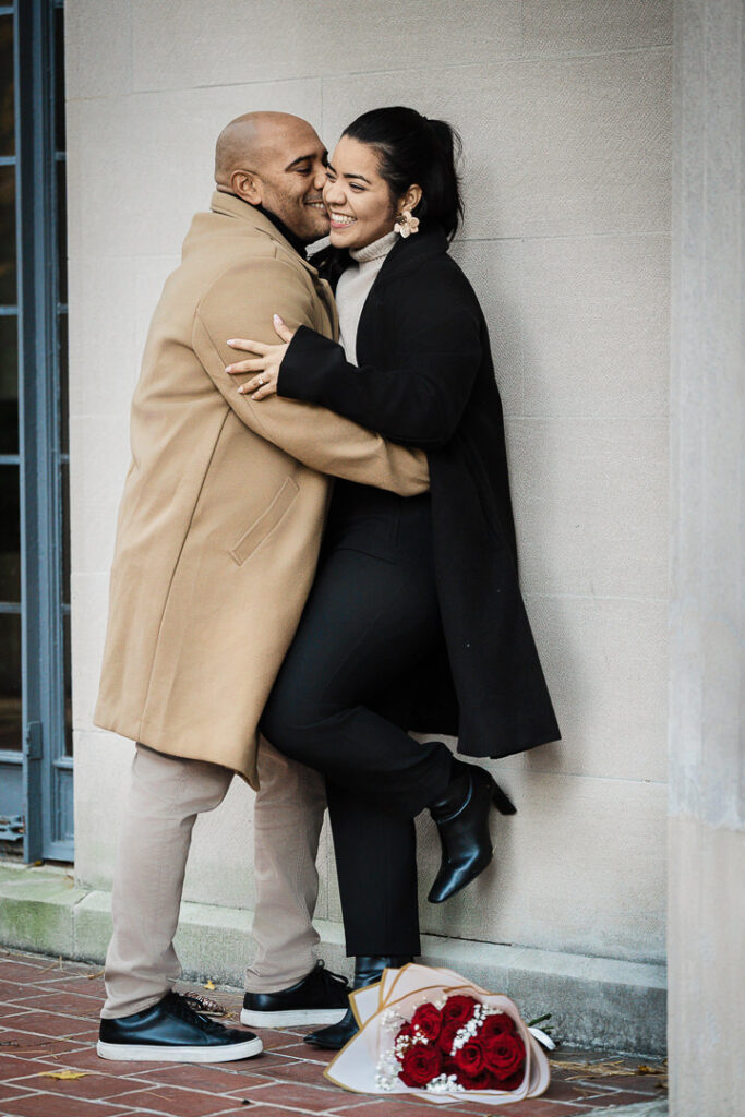 Newly engaged couple embraces against Van Vleck garden wall with red roses on ground, romantic proposal photography Montclair NJ