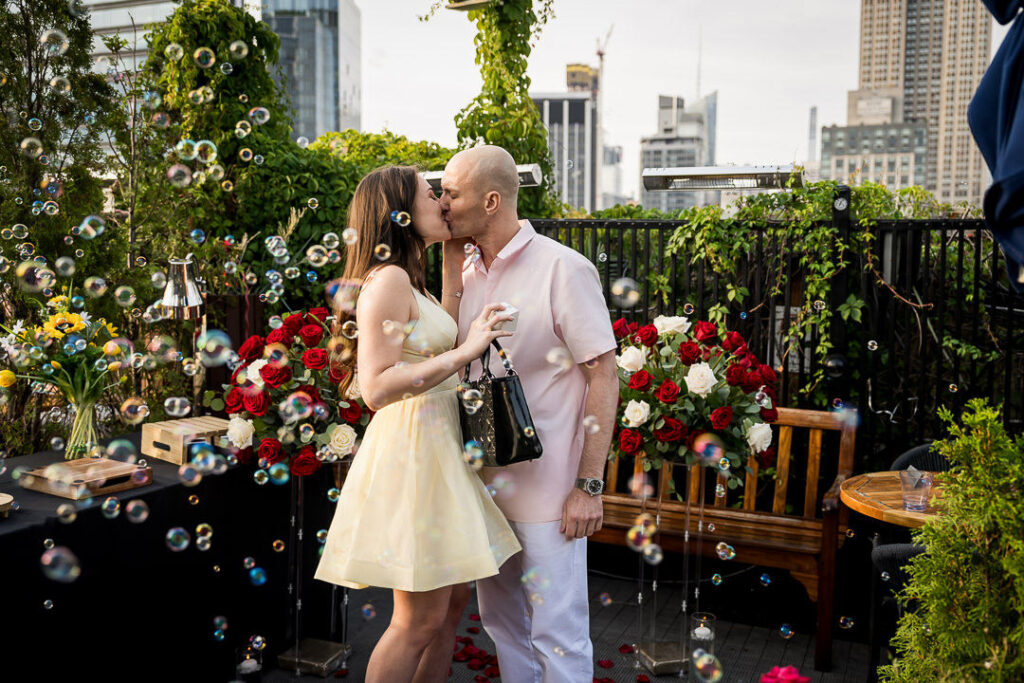 Engaged couple kisses surrounded by floating bubbles with Empire State Building and Manhattan skyline backdrop, professional engagement photography Northern New Jersey