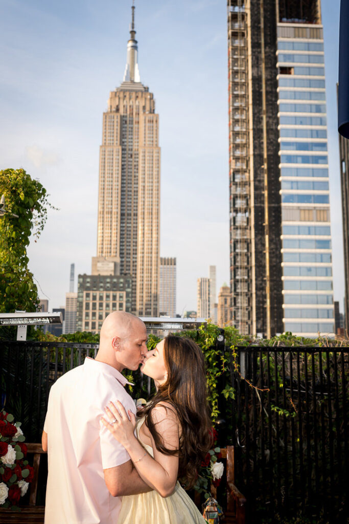 Engaged couple shares intimate kiss with Empire State Building prominently featured in Manhattan skyline background, professional proposal photography