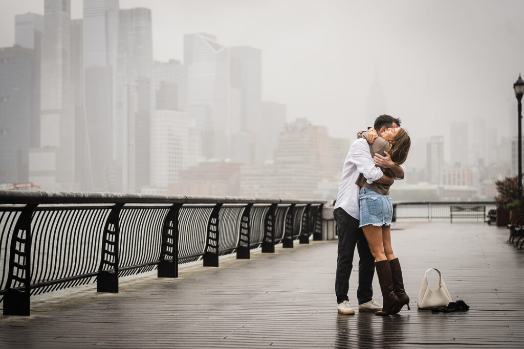 Engaged couple in romantic embrace with foggy Manhattan skyline visible from Pier 13 Hoboken