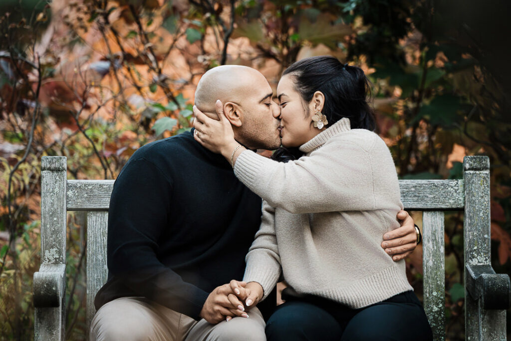 Newly engaged couple shares intimate kiss on Van Vleck garden bench with autumn foliage background, romantic engagement photography Montclair NJ